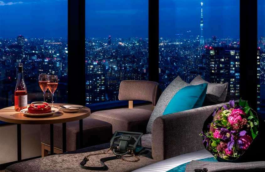 Sala de um hotel com sofás, poltronas, mesa com bolo e vinhos, buquê de flores e janela com paisagem da cidade