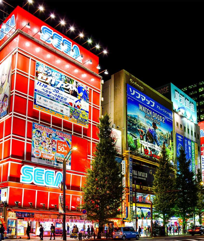 Durante a noite, uma das ruas para conhecer tokyo com prédios, árvores, pessoas e carros ao redor