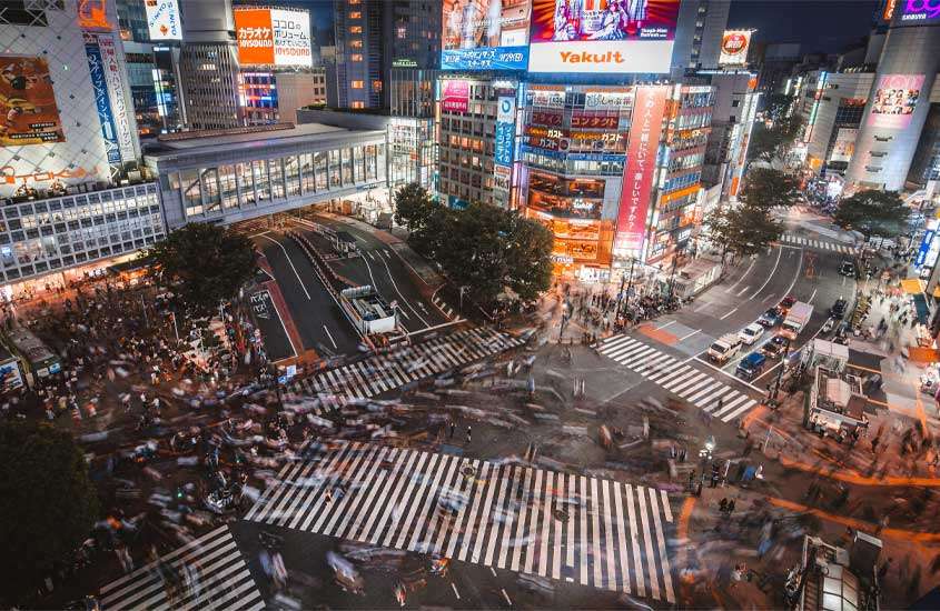 Durante a noite, cruzamento de shibuya com com diversos prédios, árvores, carros e pessoas ao redor