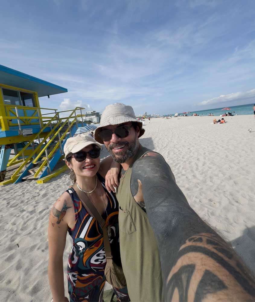 Em um dia de sol, Bárbara Rocha Alcantelado e Vagner Alcantelado em uma praia de Miami com pessoas ao redor