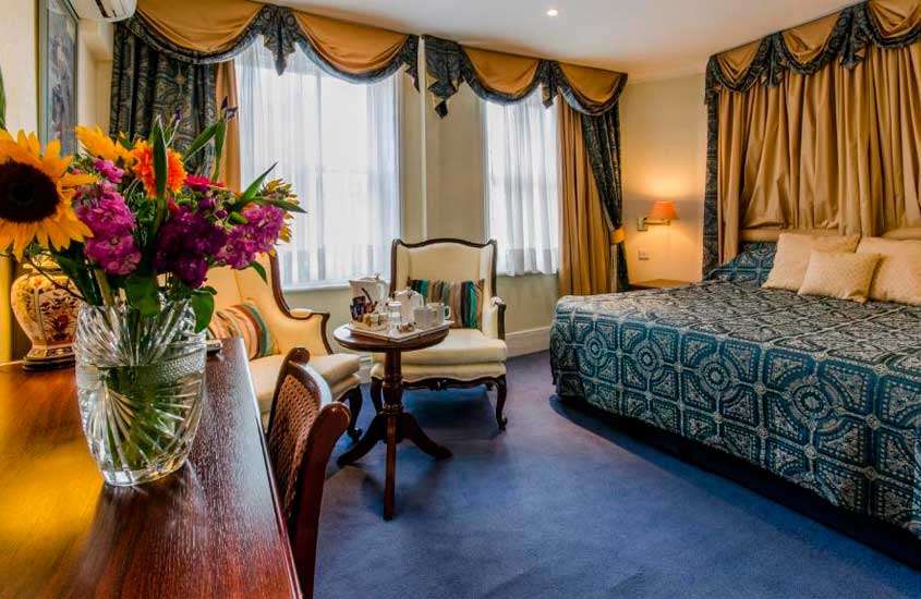 Suíte luxuosa de um hotel com cama grande de dossel, poltronas, flores, cadeira e janelas grandes acortinadas