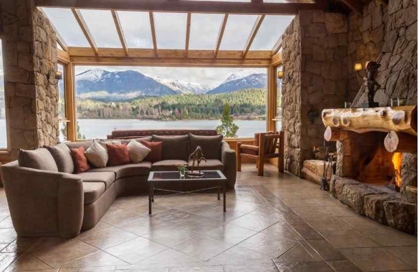 Em um dia nublado, interior de um dos melhores hotéis em Bariloche para família com sofá, lareira, rio do lado e montanhas atrás