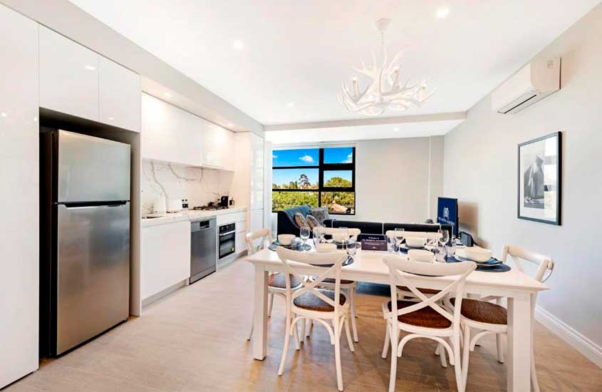 Sala com cozinha conjugada de um hotel onde ficar em Melbourne, com mesa, cadeiras, eletrodomésticos modernos, sofás, janela, TV e quadro decorativo