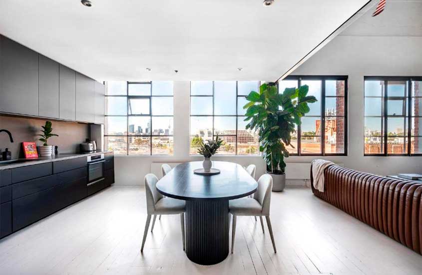 Sala com cozinha cojugada de um hotel onde ficar em Melbourne, com mesa, cadeiras, armários, sofá, plantas decorativas e janelas grandess