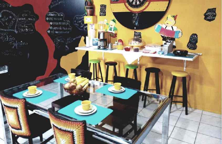 Área de café da manhã do hotel com mesas, cadeiras, bancos e decorações coloridas