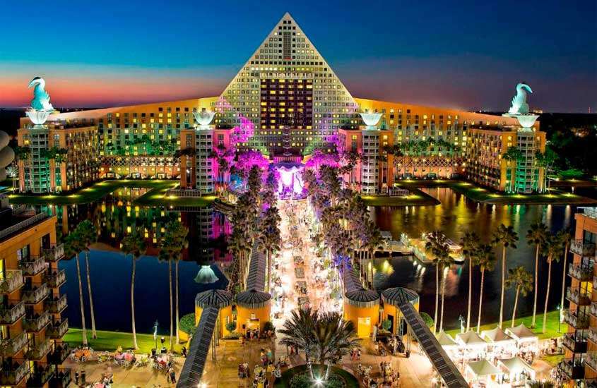 Durante o anoitecer, visão aérea de um dos hotéis para passa o Réveillon na Disney, com árvores, lagos,luzes coloridas e pessoas ao redor