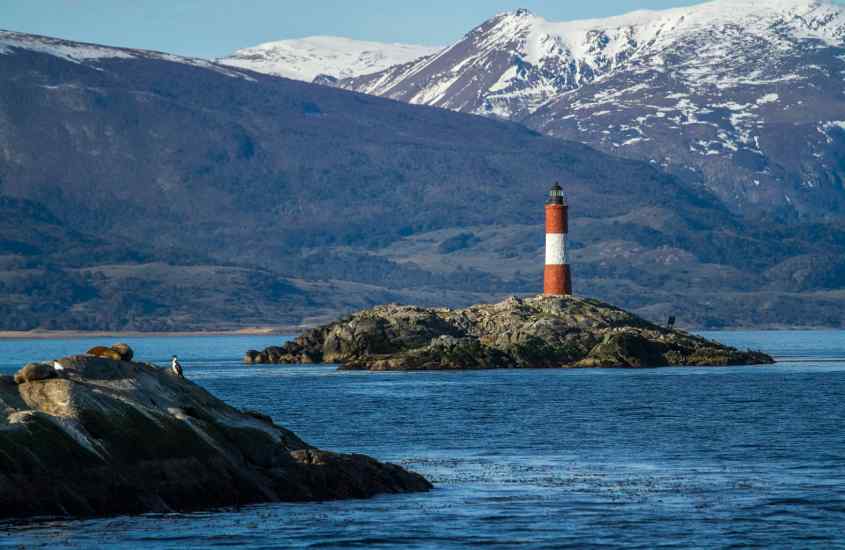 Em um dia de sol, mar de Ushuaia com montanhas ao redor e farol no meio, uma das cidades para passar o réveillon na Argentina