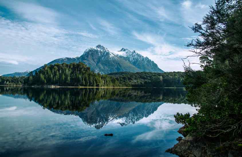 Em um dia de sol, lago em Bariloche com árvores e montanhas ao redor, uma das cidades para passar o réveillon na Argentina