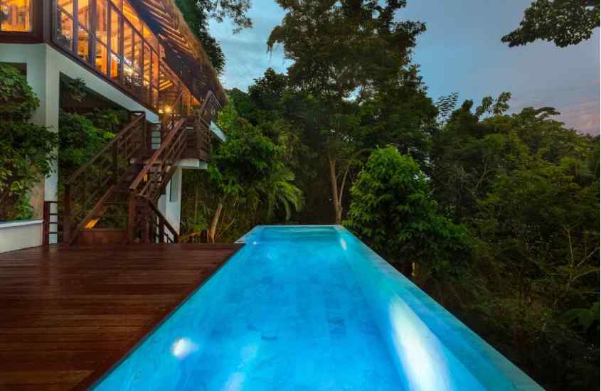 Durante o anoitecer, área de lazer de um hotel com piscina, deck de madeira, árvores e plantas ao redor