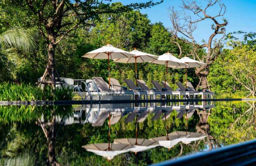 Em um dia de sol, área de lazer de hotel com piscina, espreguiçadeiras, guarda-sóis e árvores ao redor, uma das opções de hotel para se hospedar durante o Réveillon na Tailândia