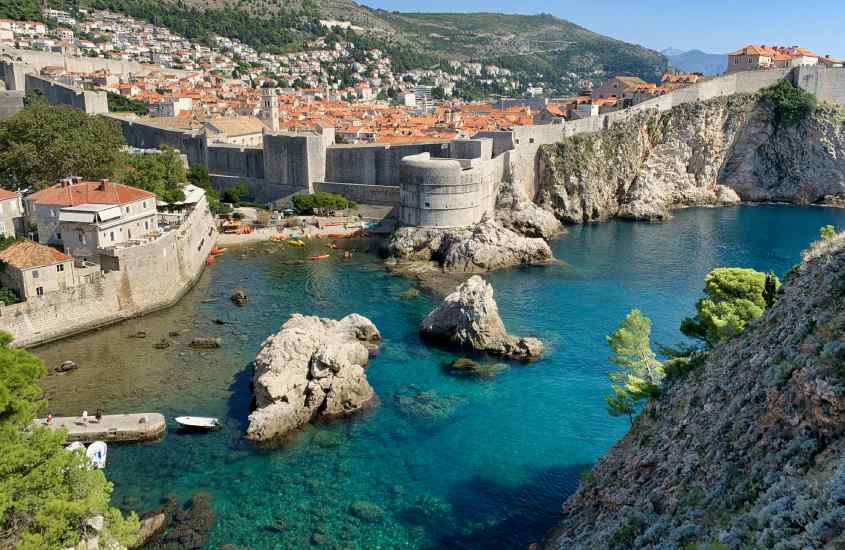 Em um dia de sol, paisagem da cidade velha, um dos bairros onde ficar em Dubrovnik com praia, paredões de pedra, casas e árvores ao redor