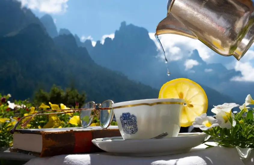 Xícara de chá com fatia de limão, livro e óculos sobre a mesa, com montanhas das Dolomitas ao fundo