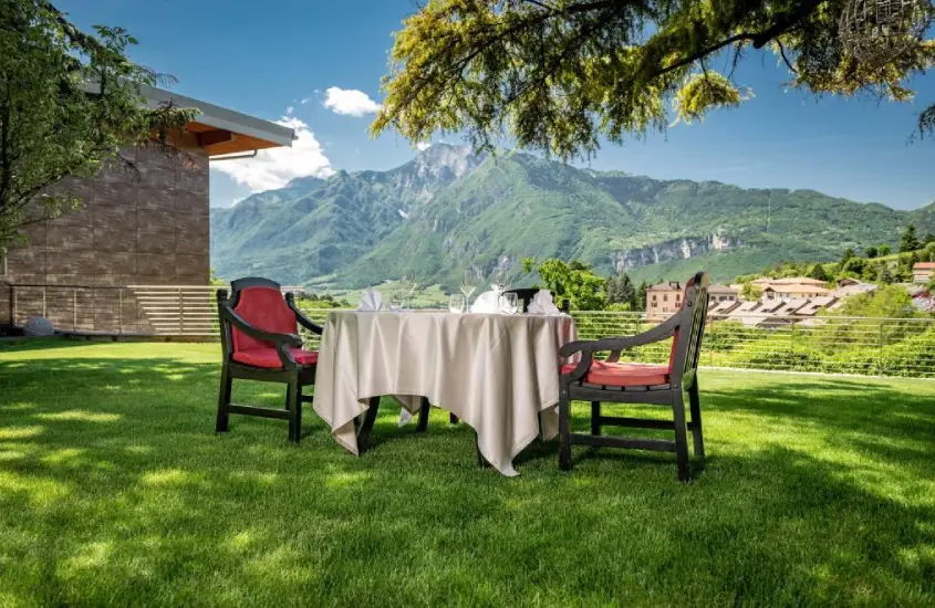 Mesa de jantar ao ar livre com toalha branca, cadeiras vermelhas e vista para montanhas verdes em Trento