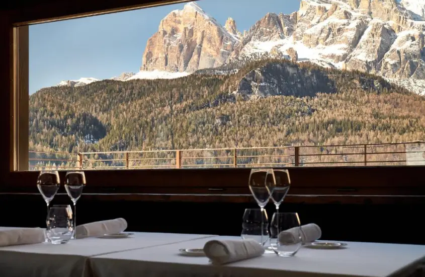 Vista das montanhas nevadas ao entardecer através de uma janela com taças de vinho na mesa