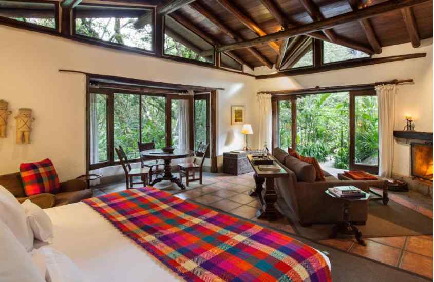 Quarto de um hotel onde se hospedar em machu picchu com cama de casal, mesas, cadeiras, lareira, sofás, janelas grandes, tapetes e varanda do lado