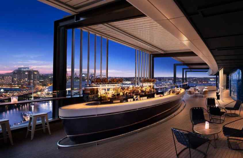 Durante o entardecer, bar e restaurante de um dos hotéis onde passar reveillon em Sydney com mesas, cadeiras e janelas com vista da cidade