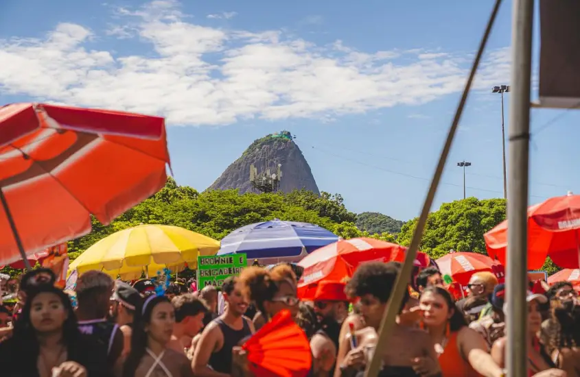 Foliões desfrutam do Carnaval no Rio de Janeiro, com o Pão de Açúcar ao fundo, sob um céu azul e coloridos guarda-sóis