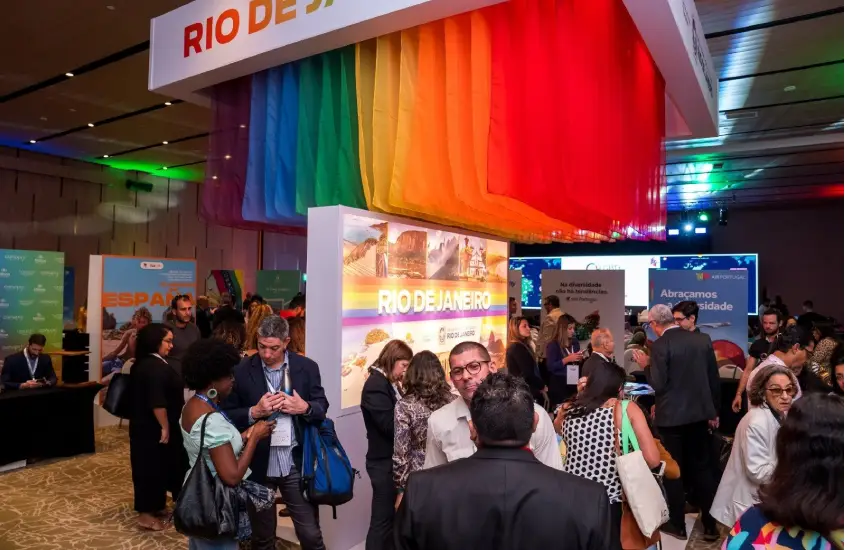 Expositores e visitantes interagem na LGBT+ Turismo Expo, com um estande do Rio de Janeiro ao fundo decorado com bandeiras coloridas