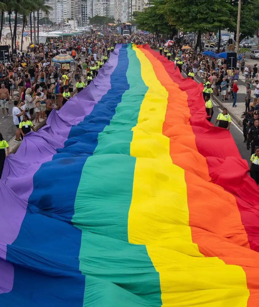 Multidão celebra a Parada do Orgulho LGBT em Copacabana com uma enorme bandeira arco-íris, rodeada por prédios e palmeiras