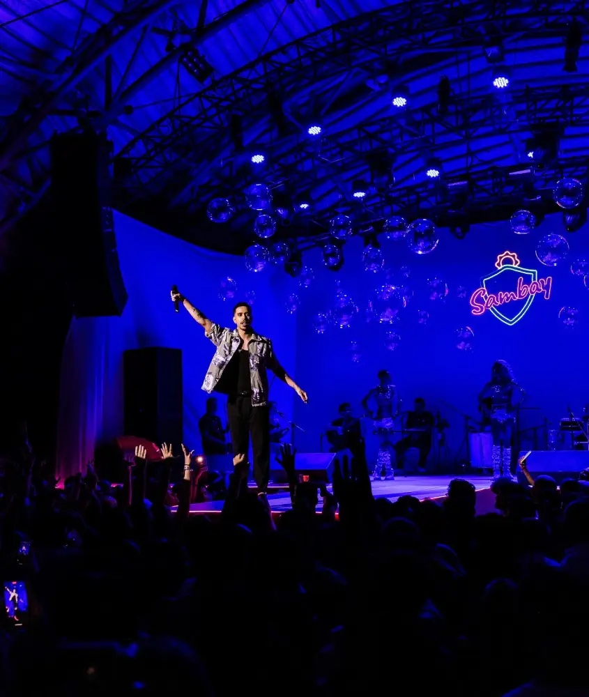 Festa Roda Sambay ao ar livre, com um palco iluminado e artistas performando para uma plateia animada