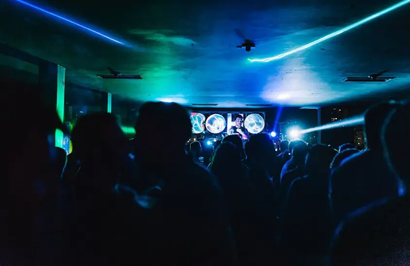 Pessoas dançando em um ambiente escuro com luzes azuis e verdes na Festa BOHO, enquanto um DJ toca música ao fundo