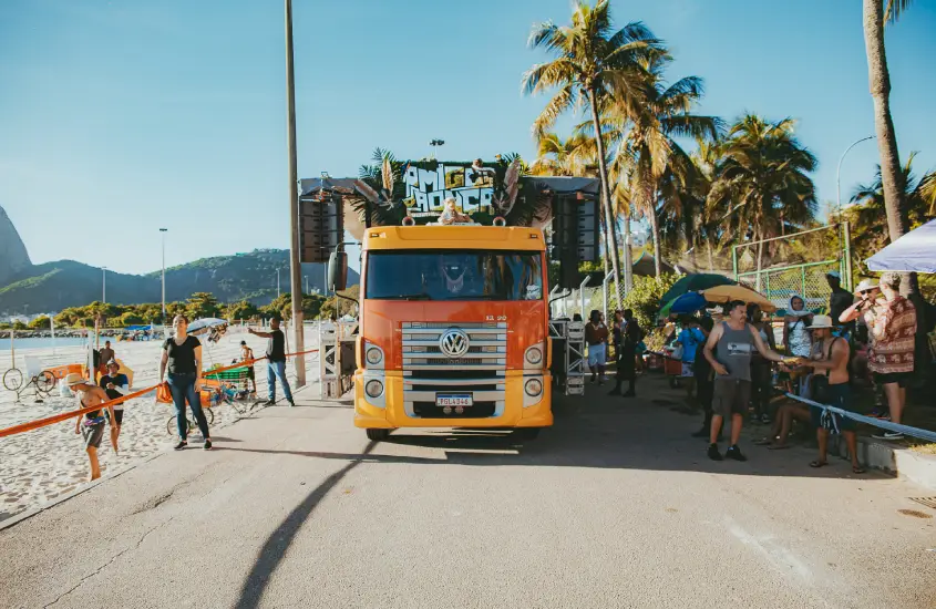 Caminhão decorado lidera o Bloco Amigos da Onça em uma avenida próxima à praia, com pessoas e palmeiras ao redor