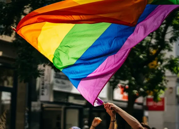 Bandeira arco-íris do orgulho LGBT sendo agitada por participantes em um evento ao ar livre, com árvores e prédios ao fundo