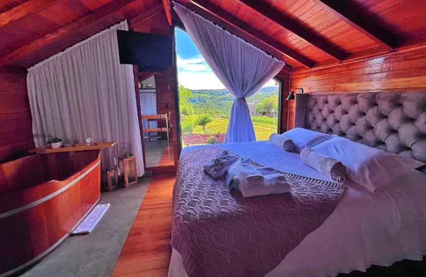 Quarto de um chalé de madeira com cama de casal, banheira, TV e janelas com paisagem das vinícolas