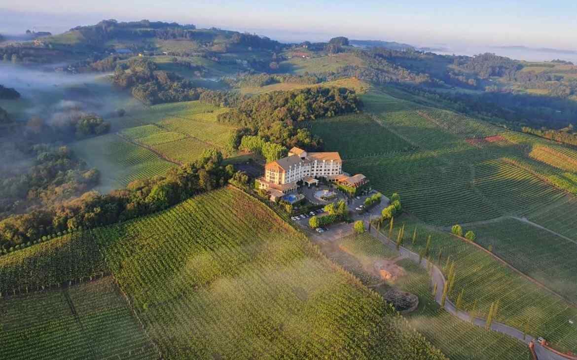 Em um dia de sol, visão aérea de um dos hotéis vinícolas no Vale dos Vinhedos com árvores e partes gramadas ao redor