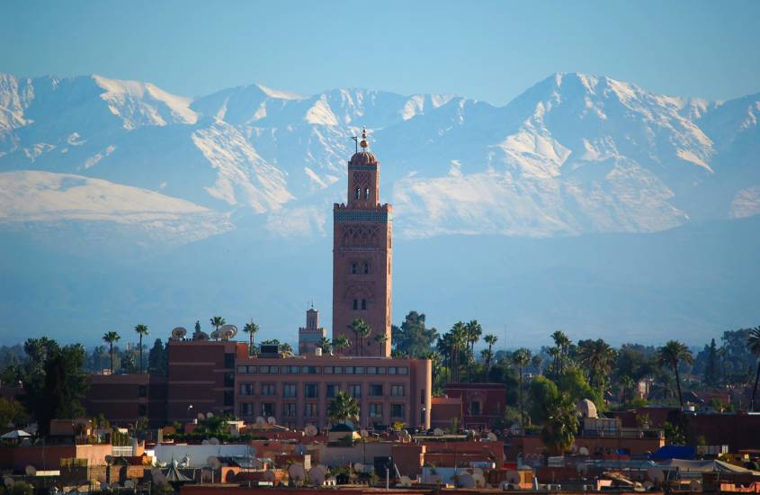 Em um dia de sol, paisagem de Marrakech com prédios, árvores ao redor e montanhas atrás