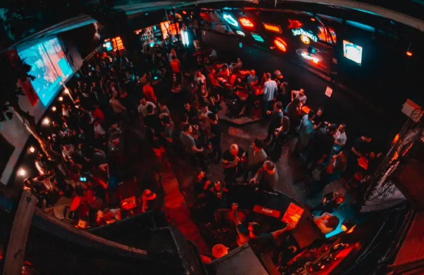 Uma vista aérea de um bar lotado com luzes neon, várias pessoas se divertindo, e mesas espalhadas pelo ambiente