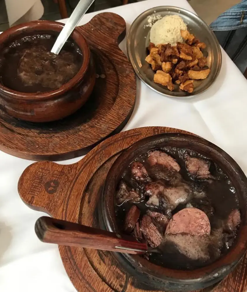 Dois pratos de feijoada servidos em panelas de barro, acompanhados de torresmo e farofa em um prato separado