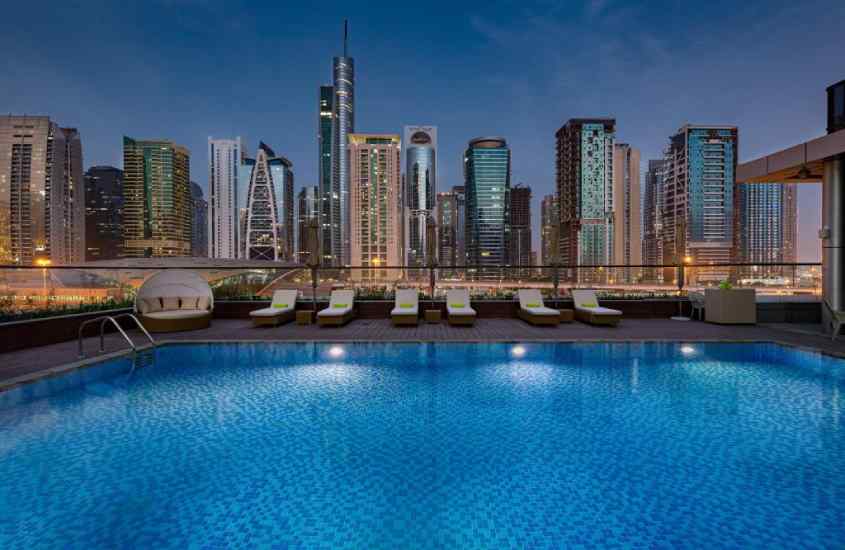 Durante a noite, área de lazer de um dos hotéis onde passar reveillon em Dubai, com piscina, espreguiçadeiras, sofás e paisagem da cidade