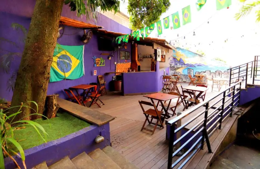 Área externa do Pura Vida Hostel com mesas, cadeiras e bandeiras do Brasil decorando o ambiente durante o dia