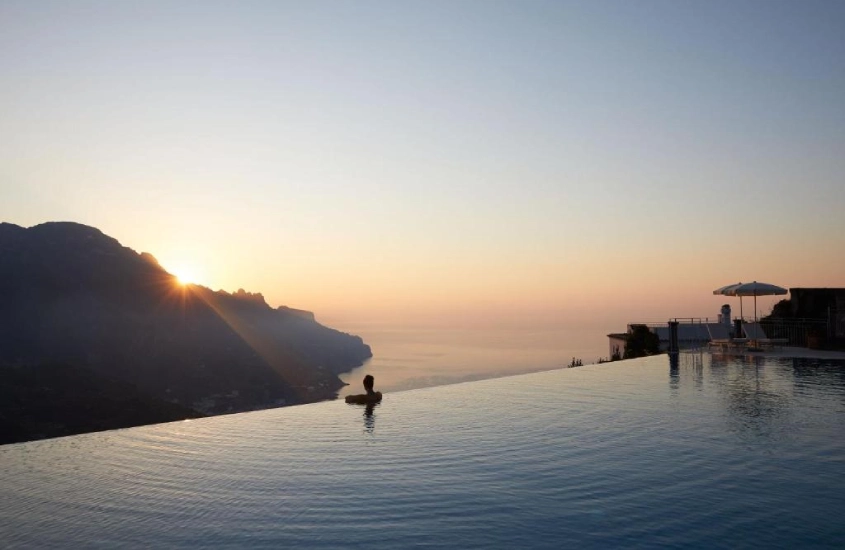 Pessoa relaxando em uma piscina infinita com vista para montanhas e o mar ao pôr do sol