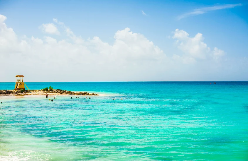 Uma praia de areia branca e mar azul-turquesa em Barbados, com palmeiras ao fundo e céu parcialmente nublado