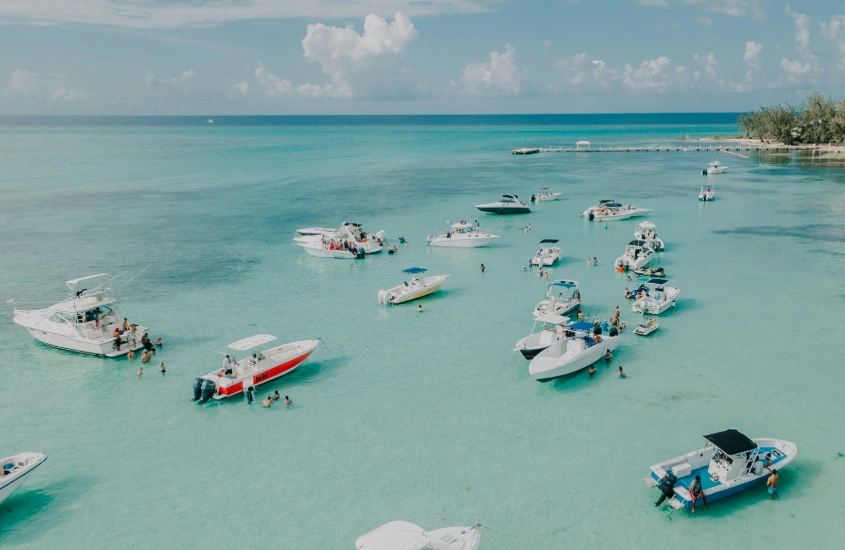 Uma baía tranquila nas Ilhas Cayman com barcos de vários tamanhos flutuando sobre a água turquesa clara, em um dia ensolarado