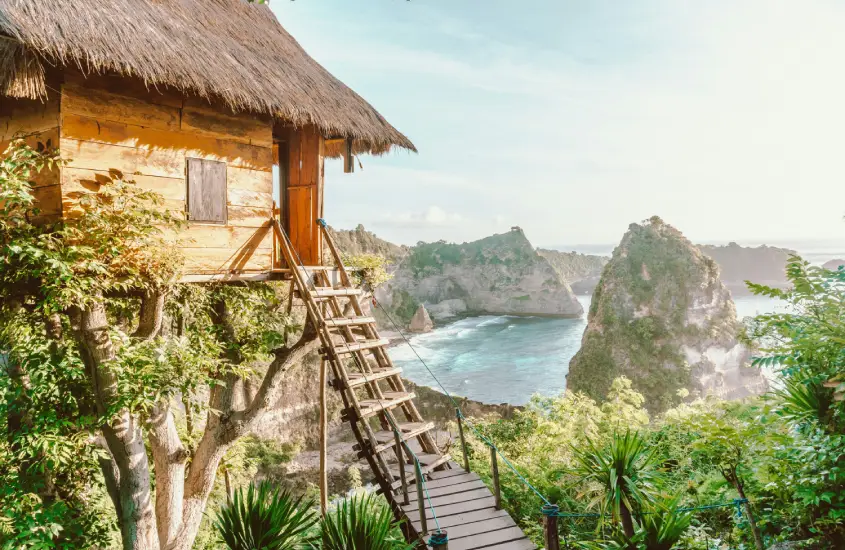 Casa de madeira rústica suspensa em uma árvore em Bali, com uma escada de madeira e uma vista deslumbrante do mar e das formações rochosas ao longe