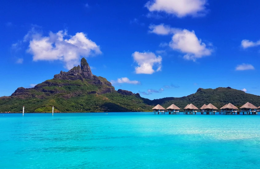 O icônico Monte Otemanu em Bora Bora, cercado por bangalôs sobre a água e o mar azul vibrante, com um céu azul brilhante e poucas nuvens