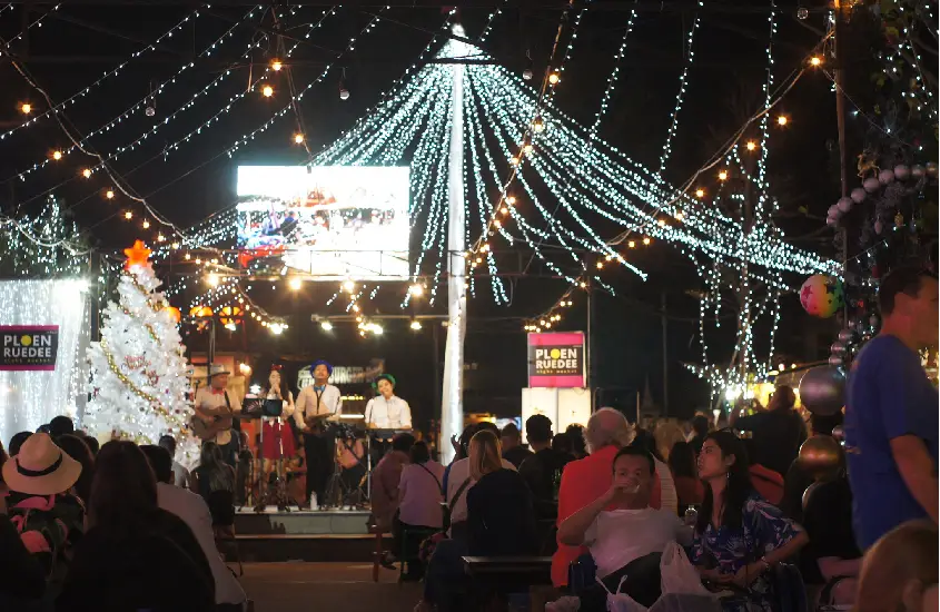 Cena noturna festiva no Ploen Ruedee Night Market, com luzes brilhantes, decoração natalina e uma banda ao vivo tocando para o público