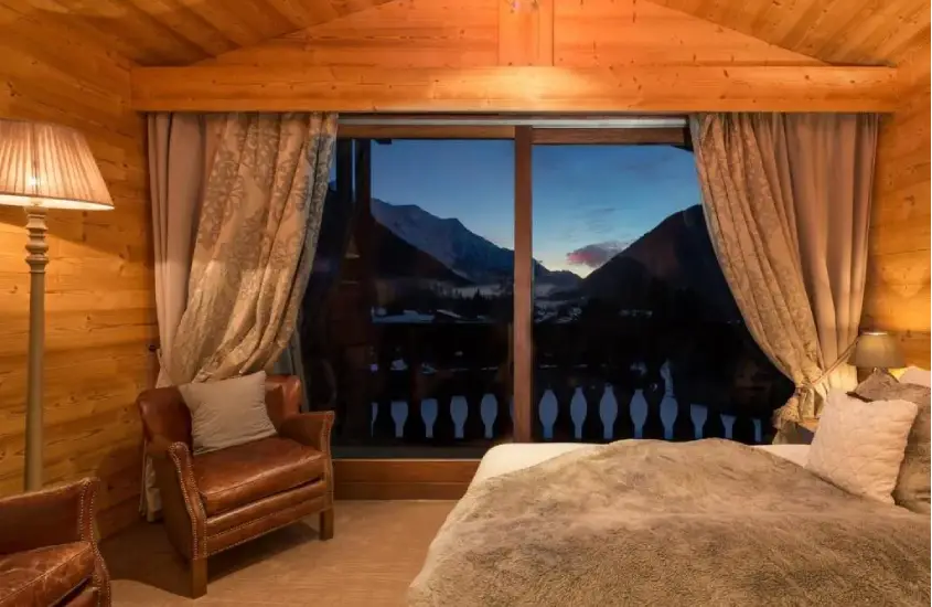 Quarto de hotel em Chamonix com decoração de madeira, poltronas de couro e uma grande janela com vista para as montanhas ao anoitecer