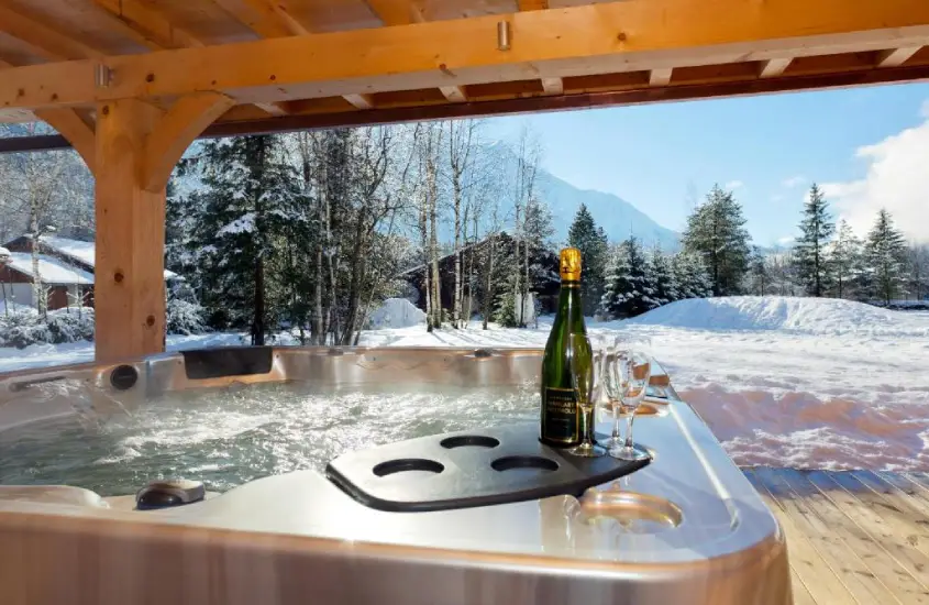 Jacuzzi coberta com vista para a neve e as montanhas ao fundo, com uma garrafa de champanhe e taças ao lado