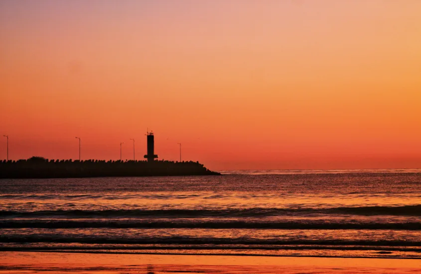 Farol de Itajaí ao amanhecer, com o mar sereno e céu alaranjado ao fundo