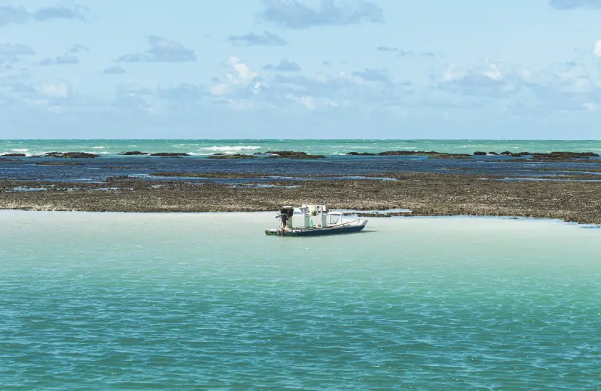 -Vista do mar cristalino com uma pequena embarcação ao centro, cercado por águas verde-claras e recifes de corais ao fundo