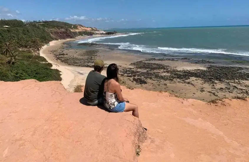 Casal sentado em um penhasco apreciando a vista panorâmica do mar azul e da praia abaixo, sob um céu limpo