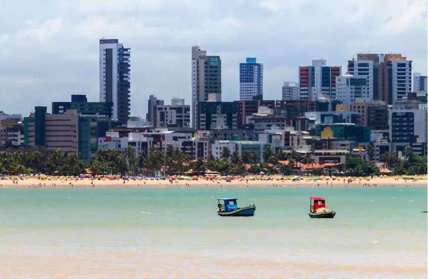 Vista panorâmica da cidade ao fundo com prédios altos e a praia movimentada com barcos coloridos na água em primeiro plano