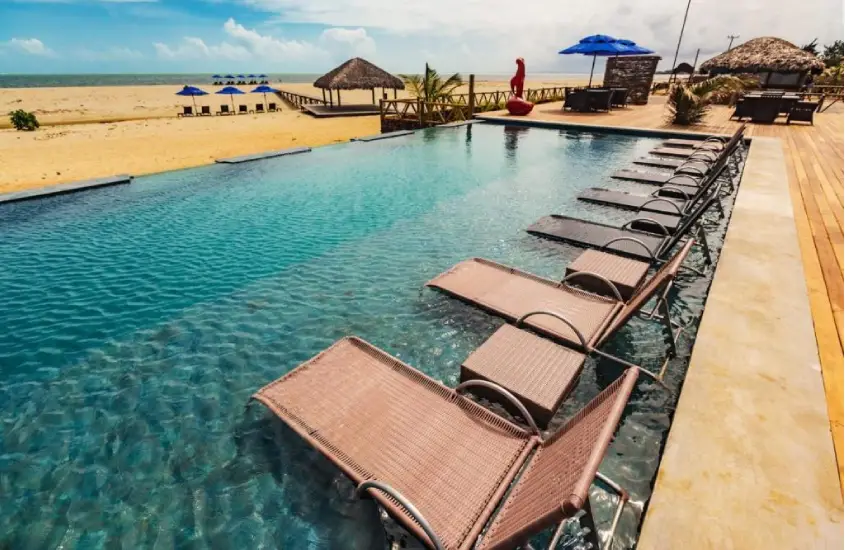 Piscina de borda infinita à beira da praia com espreguiçadeiras dentro da água e guarda-sóis ao fundo