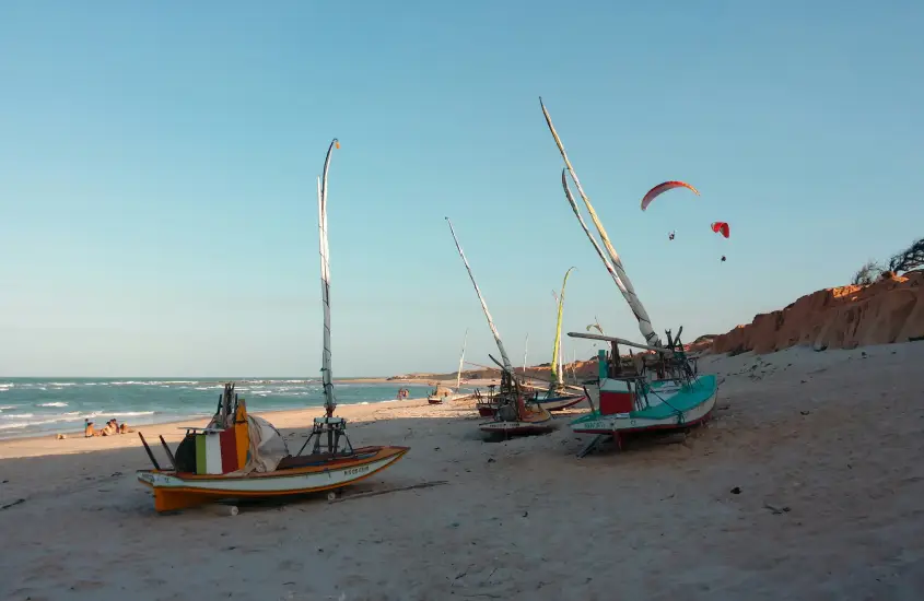 Várias jangadas coloridas na areia da praia durante o dia, com pessoas ao fundo e parapentes no céu