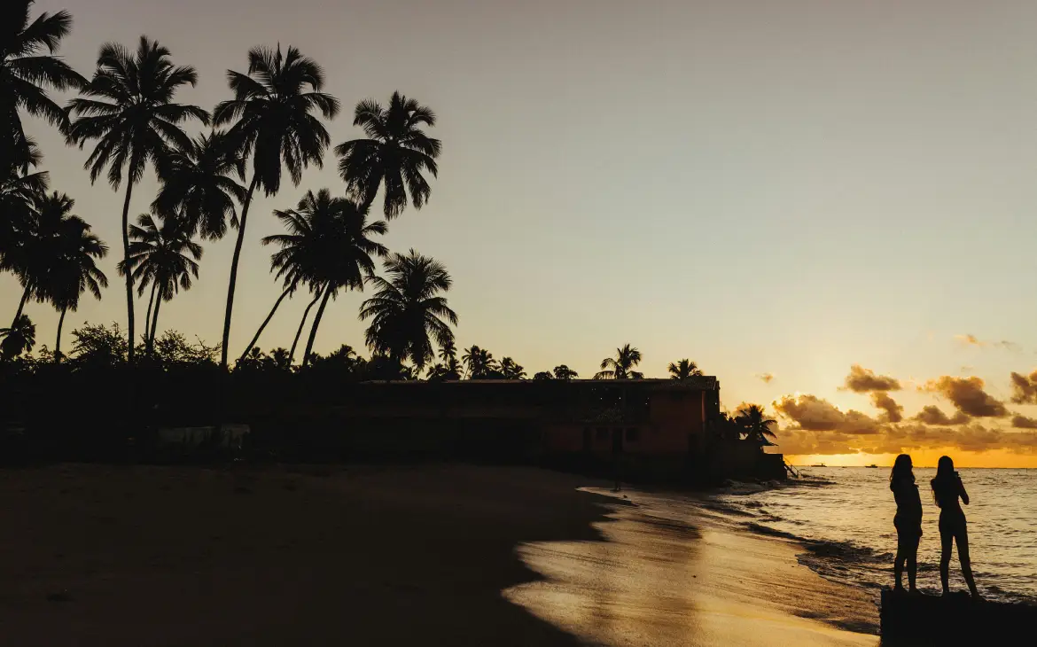 Pôr do sol em uma praia com coqueiros silhuetados e duas pessoas apreciando a vista