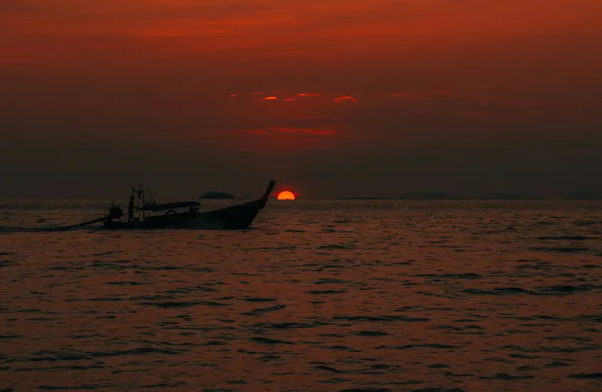 Longtail boat no mar durante o pôr do sol, com o céu alaranjado e o sol se pondo no horizonte em Koh Phi Phi.